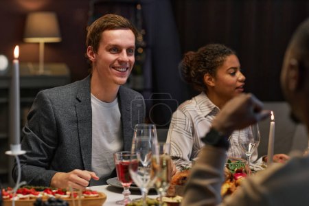 Foto de Joven feliz mirando a su amigo durante la conversación por la cena festiva mientras está sentado frente a él por la mesa servida en la fiesta en casa - Imagen libre de derechos