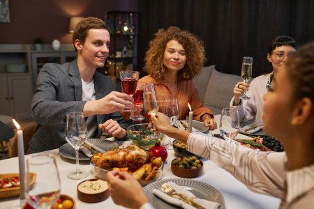 Foto de Grupo de jóvenes felices tintineando con bebidas alcohólicas sobre la mesa servida mientras cenan en casa y disfrutan de la fiesta - Imagen libre de derechos