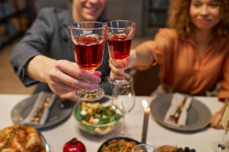 Photo for Focus on two glasses of red wine held by young cheering man and woman sitting by table served with homemade food prepared for guests - Royalty Free Image