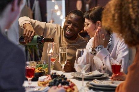 Foto de Feliz joven afroamericano hombre vertiendo vino tinto en la flauta de su novia mientras se sienta junto a ella por la mesa servida en la fiesta en casa - Imagen libre de derechos