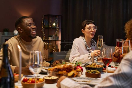 Foto de Feliz joven afroamericano riendo mientras está sentado junto a la mesa servida junto a su novia con flauta de champán durante la cena - Imagen libre de derechos