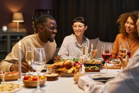 Foto de Riendo hombre negro con flauta de champán inclinado sobre la mesa servida durante el chat con sus amigos mientras disfruta de la fiesta en casa y la cena - Imagen libre de derechos