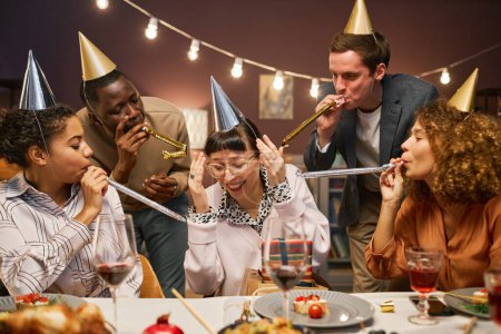 Foto de Grupo de jóvenes interculturales en gorras de cumpleaños soplando a los fabricantes de ruido en los oídos de la chica morena sonriente mientras están sentados junto a la mesa servida juntos - Imagen libre de derechos