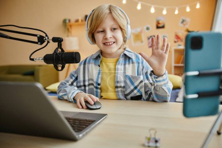Photo for Smiling boy waving hand to online audience and speaking in microphone while making livestream in front of laptop and smartphone by desk - Royalty Free Image