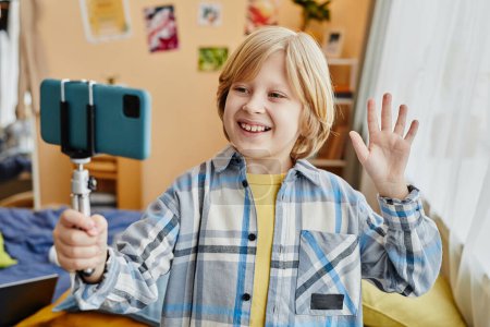Blond smiling schoolboy and blogger waving hand to online audience while making livestream and looking at smartphone screen