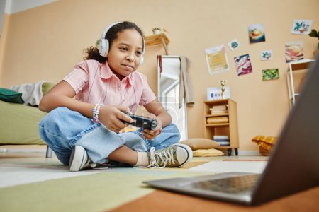 Photo for Happy schoolgirl with joystick pressing buttons and looking at laptop screen while sitting on the floor and playing video game at home - Royalty Free Image
