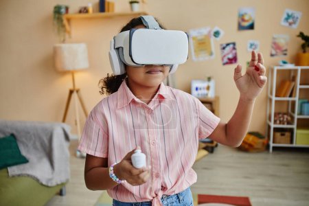 Téléchargez les photos : Mignon AFrican Américaine écolière en VR casque jouer jeu virtuel tout en se tenant devant la caméra et en appuyant sur le bouton sur la manette de jeu - en image libre de droit