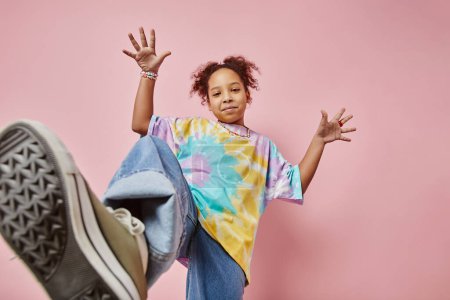 Foto de Feliz chica juguetona en ropa de casualwear posando delante de la cámara en el fondo rosa, mientras que de pie por la pared durante la sesión de fotos - Imagen libre de derechos