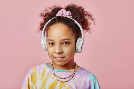 Photo for African American schoolchild in headphones and t-shirt looking at camera while standing against pink background with copyspace around - Royalty Free Image