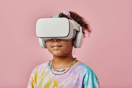 Téléchargez les photos : Écolière avec casque VR debout devant la caméra sur fond rose pendant le jeu vidéo tout en posant en isolement - en image libre de droit