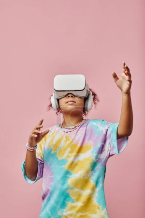 Photo for Modern schoolgirl in t-shirt and VR headset reaching to virtual object while playing three dimension video game against pink background - Royalty Free Image