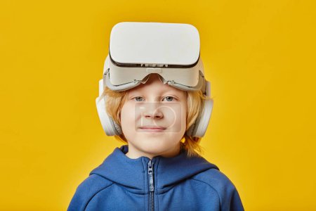Mignon garçon blond avec casque VR sur le front en regardant la caméra après avoir joué au jeu virtuel tout en se tenant sur fond jaune
