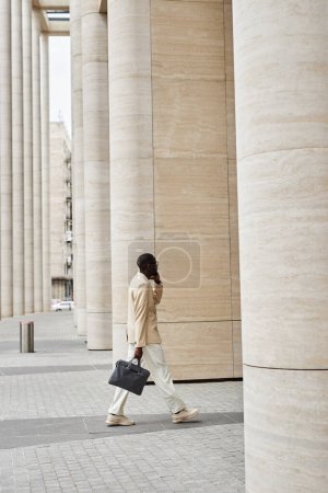Seitenansicht eines jungen eleganten männlichen Unternehmers im eleganten weißen Anzug, der auf einem Mobiltelefon spricht, während er sich in Richtung eines riesigen modernen Gebäudes bewegt