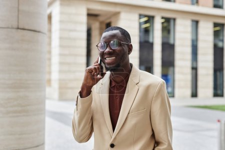 Junger erfolgreicher afroamerikanischer Geschäftsmann in Elfenbeinjacke spricht auf Smartphone, während er in urbaner Umgebung vor der Kamera steht