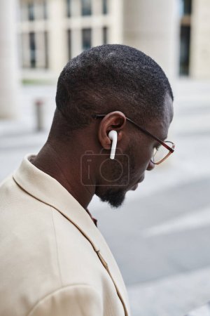 Seitenansicht eines jungen afroamerikanischen Geschäftsmannes mit Brille und Kopfhörer, der im Freien auf den Bildschirm eines mobilen Geräts blickt