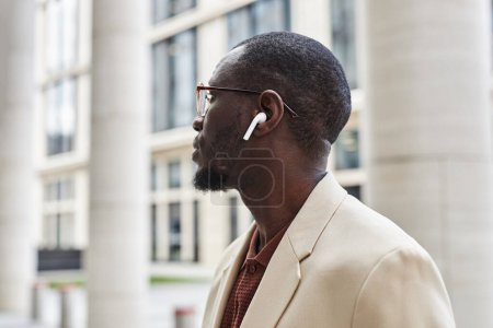 Seitenansicht eines jungen, gelassenen schwarzen Mannes im eleganten weißen Anzug, Brille und Kopfhörer, der Musik hört und etwas Kurioses betrachtet
