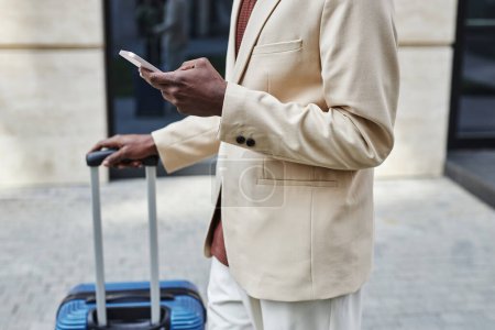Schnappschuss eines jungen afroamerikanischen Geschäftsmannes mit Koffer und Smartphone, während er vor der Kamera vor einem modernen Gebäude steht