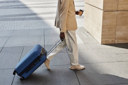 Foto de Foto recortada de hombre de negocios moderno con taza de café y maleta con equipaje caminando por la calle mientras se apresura al aeropuerto o estación de autobuses - Imagen libre de derechos