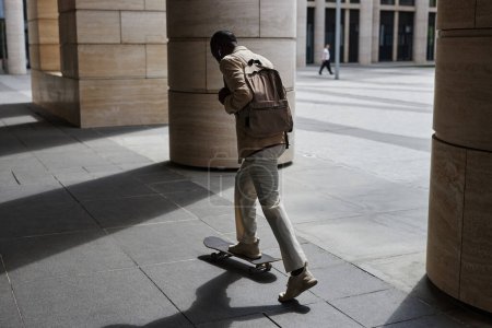 Rückansicht eines jungen Geschäftsmannes mit Rucksack, der auf einem Skateboard steht, während er an riesigen Säulen des modernen Gebäudes entlang zur Arbeit fährt