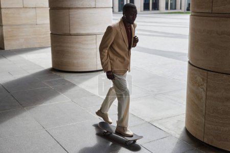 Junger afroamerikanischer Angestellter in ruhiger Luxuskleidung fährt Skateboard an Säulen moderner Gebäude entlang, während er zur Arbeit eilt