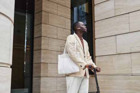 Junger moderner Geschäftsmann in eleganter Kleidung, der an Rollergriffen hält, während er am Gebäude steht und Musik über Kopfhörer hört