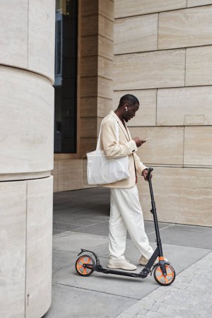 Foto de Largo tiro de empresario afroamericano con scooter, smartphone y bolso textil blanco de pie por la arquitectura moderna y mensajes de texto - Imagen libre de derechos