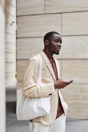 Foto de Joven hombre de negocios sereno con bolso textil blanco en el hombro esperando a su colega y utilizando el teléfono inteligente mientras está de pie delante de la cámara - Imagen libre de derechos
