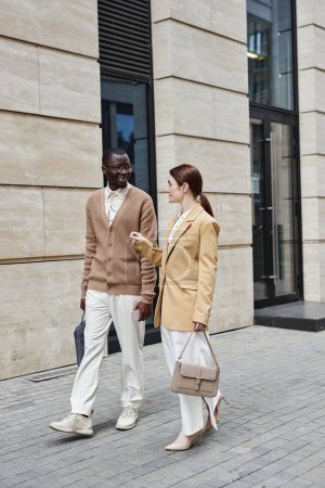 Lange Aufnahme von zwei jungen interkulturellen Managern in ruhiger Luxuskleidung, die nach der Arbeit den Bürgersteig entlanggehen und über Arbeitspunkte diskutieren