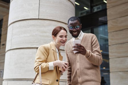 Two young cheerful colleagues in quiet luxury apparel looking at screen of smartphone held by smiling African American male employee