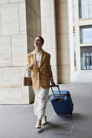 Junge hübsche Geschäftsfrau in ruhiger Luxuskleidung zieht Koffer mit Gepäck und trägt Handtasche über dem Arm, während sie zum Flughafen eilt