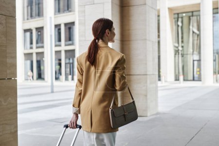 Rückansicht einer jungen brünetten Geschäftsfrau in Formalwear, die Koffer zieht, während sie zum Flughafen oder Busbahnhof entlang moderner Architektur läuft