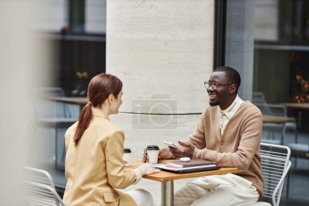 Téléchargez les photos : Heureux jeune homme d'affaires afro-américain avec smartphone parlant à une collègue brune alors qu'elle était assise devant elle dans un café de rue - en image libre de droit