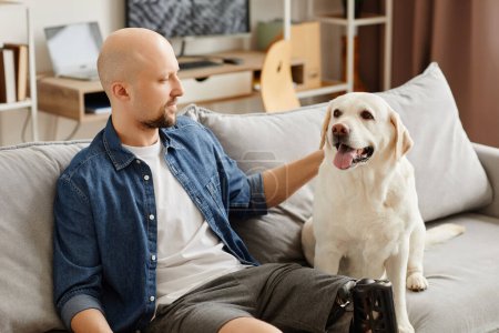 Portrait d'homme adulte souriant avec jambe prothétique relaxant sur le canapé avec chien blanc heureux