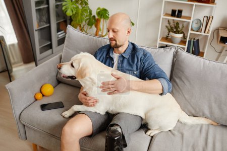 Portrait grand angle de l'homme adulte handicapé tenant grand chien blanc dans les genoux tout en se relaxant sur le canapé ensemble