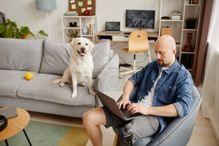 Foto de Retrato de hombre adulto con discapacidad utilizando el ordenador portátil en la sala de estar con perro blanco en el fondo, espacio de copia - Imagen libre de derechos