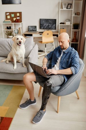 Téléchargez les photos : Portrait vertical de l'homme adulte avec prothèse de jambe en utilisant un ordinateur portable dans le salon avec chien blanc en arrière-plan - en image libre de droit
