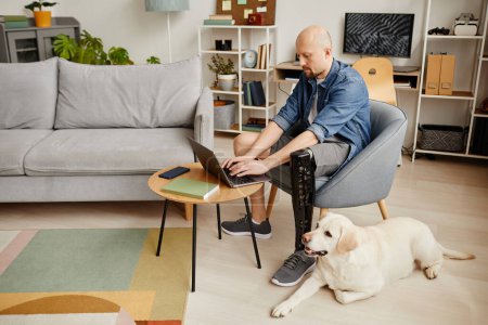 Téléchargez les photos : Portrait complet de l'homme adulte avec prothèse de jambe à l'aide d'un ordinateur portable à la maison et chien labrador caressant, espace de copie - en image libre de droit