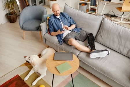 Vue du dessus de l'homme adulte avec la jambe prothétique relaxant sur le canapé dans le salon et défilement des médias sociaux, espace de copie