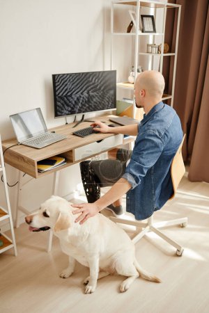 Portrait latéral de l'homme adulte handicapé à l'aide d'un ordinateur au bureau travaillant à la maison en informatique et caressant chien labrador blanc, espace de copie