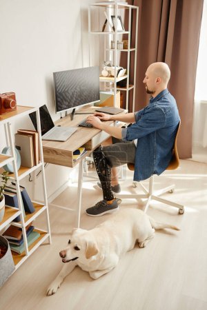 Retrato de vista lateral de hombre adulto con pierna protésica usando computadora trabajando desde casa con perro labrador esperando