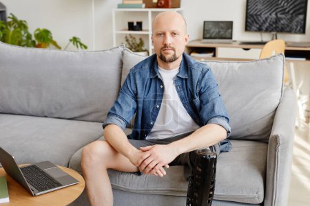 Téléchargez les photos : Portrait de l'homme adulte avec une jambe prothétique regardant la caméra alors qu'il était assis sur le canapé à la maison, espace de copie - en image libre de droit