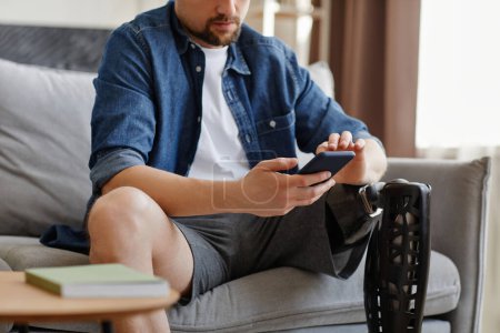 Foto de Primer plano del hombre con la pierna protésica usando el teléfono inteligente sentado en el sofá en casa, espacio de copia - Imagen libre de derechos