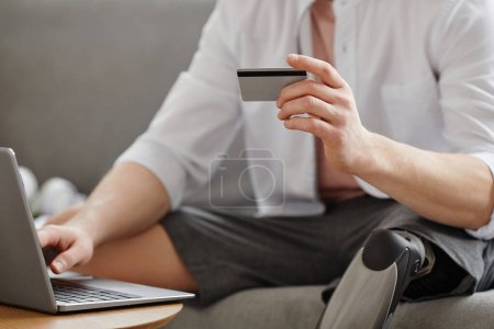 Closeup of man with prosthetic leg entering credit card information online while using laptop, copy space