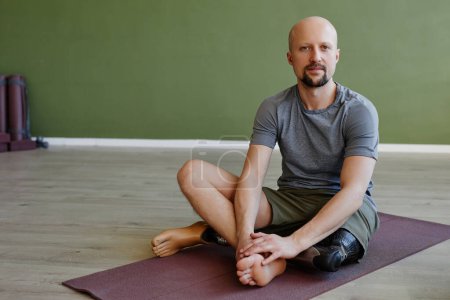 Téléchargez les photos : Portrait complet de l'homme adulte avec prothèse de jambe assis sur tapis de yoga à l'intérieur et en regardant la caméra - en image libre de droit