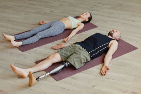 Téléchargez les photos : Jeune couple sportif avec un homme handicapé faisant une thérapie de yoga ensemble et couché en méditation - en image libre de droit