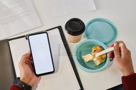 Téléchargez les photos : Gros plan d'un homme d'affaires méconnaissable tenant un smartphone avec une maquette vierge tout en mangeant des plats à emporter sur le lieu de travail, espace de copie - en image libre de droit