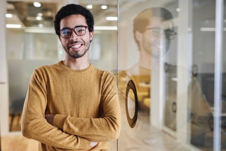 Foto de Retrato de la cintura hacia arriba del joven árabe que lleva gafas mientras está de pie junto a la pared de vidrio en la oficina y sonríe a la cámara con confianza, copia el espacio - Imagen libre de derechos