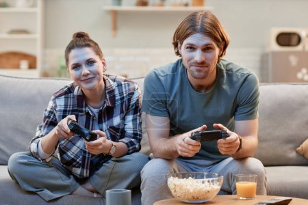 Téléchargez les photos : Portrait de jeune couple jouant ensemble à des jeux vidéo assis sur un canapé et tenant des contrôleurs - en image libre de droit