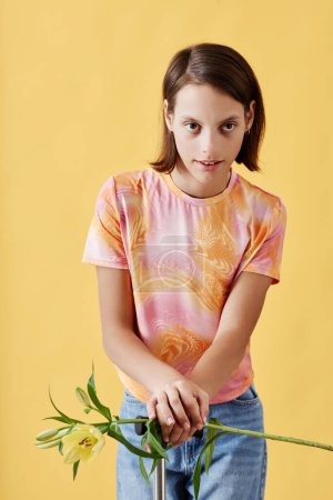 Téléchargez les photos : Portrait vertical de jeune fille avec paralysie cérébrale tenant fleur de lis et regardant la caméra debout sur fond jaune - en image libre de droit