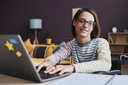 Téléchargez les photos : Portrait d'une adolescente souriante handicapée utilisant un ordinateur portable à la maison et regardant la caméra, copiez l'espace - en image libre de droit